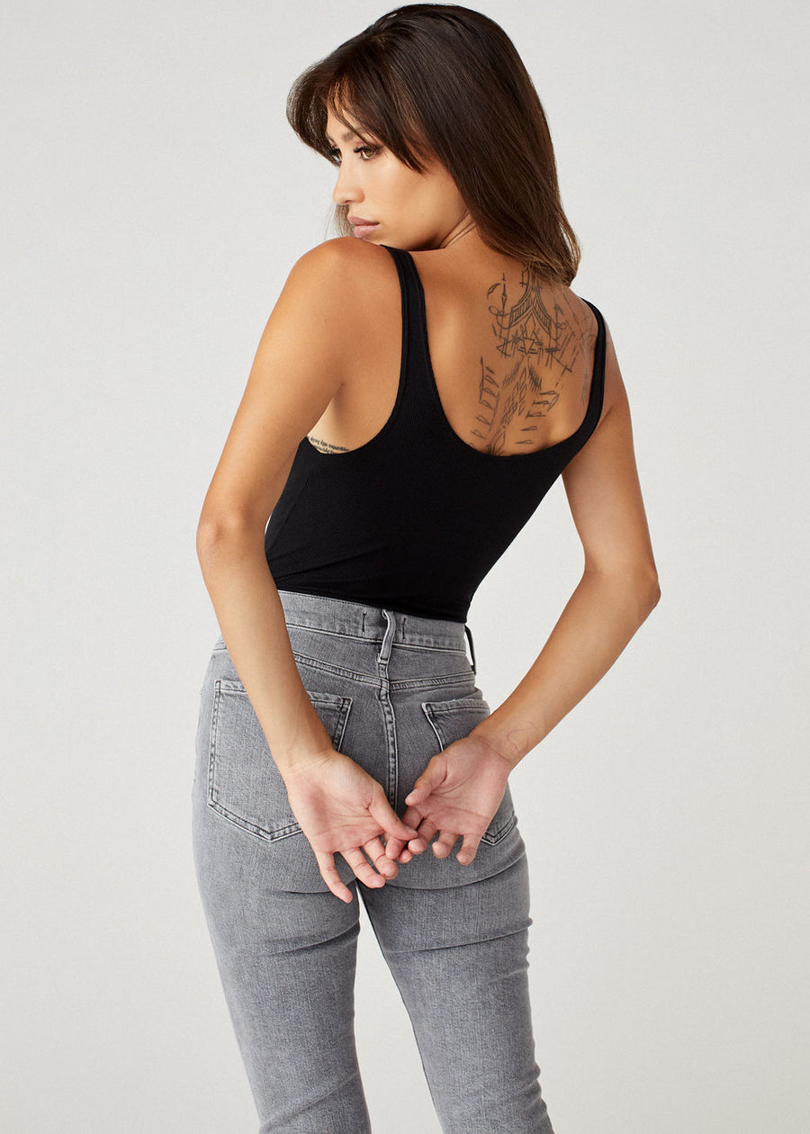 A model wearing the black Brittaney bodysuit tank top by Clyque and high waisted skinny jeans in light grey. The model is back facing the camera showing the scooped opened back.