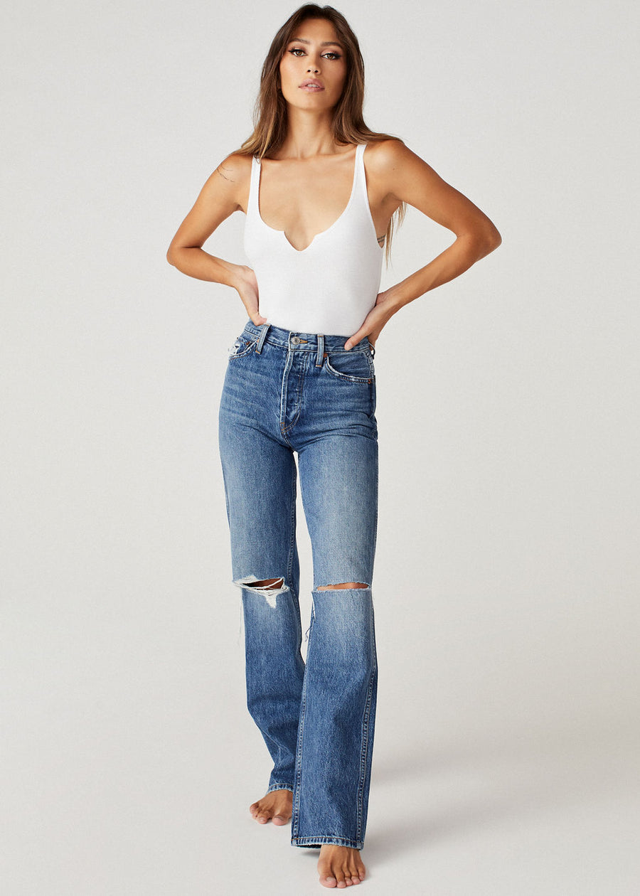 The brunette model wearing a white Brittaney strappy tank from Clyque with a V slit and blue flare crop jeans with ripped knee.